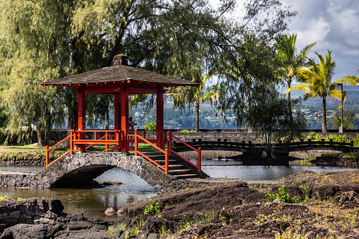 Hilo, Hawaii, USA. - January 9, 2012: Closeup of Japanese black and red small bow bridge over pond in Liliuokalani Gardens. Green trees and black rocks under blue sky with big white clouds.