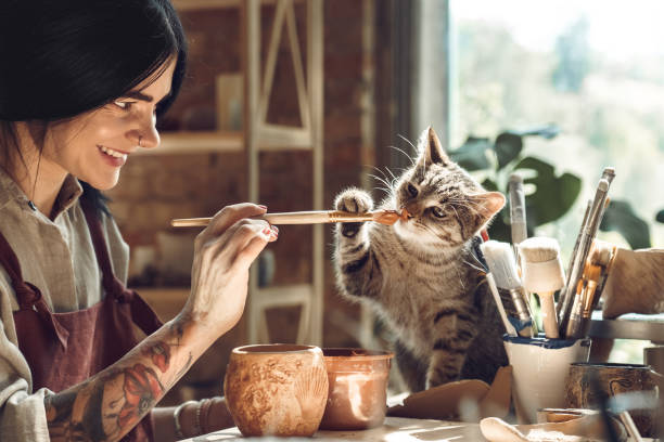 istock Craftsperson Concept. Young tattooed woman with piercing making pottery at creative studio sitting near window holding brush playing with cat laughing playful 1205627283