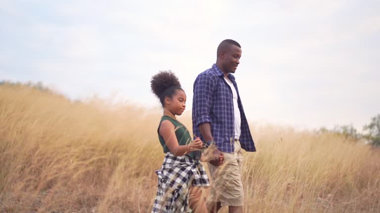 HD Slow motion Happy family African father and daughter hiking and holding hands together. Relaxing father and pretty daughter talking and walking together on golden meadow grass field in summertime