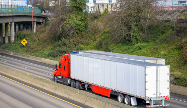 istock Big rigs semi trucks with dry van semi trailers running side by side on the wide divided multiline highway road with bridge at the city area 1205652862