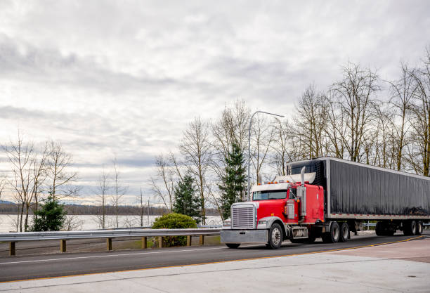 istock Big rig red classic bonnet semi truck transporting cargo in covered rubberized black semi trailer driving on the road intersection along the river 1205653781