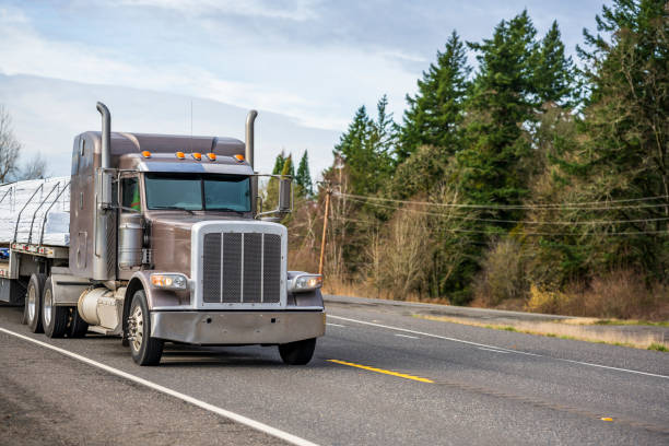 istock Powerful classic big rig brown semi truck with vertical pipes transporting packed cargo on flat bed semi trailer running on the road with marked lines 1205654521