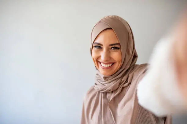 Arab beautiful woman smiling and selfie taking pictures by her mobile phone on gray backgound. Portrait of young muslim woman posing taking selfie photo with mobilephone