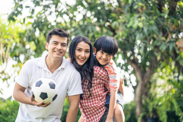 portrait of father holding soccer or football with mother and son. mom carrying son on her back with smile in garden at home. happy family activity and enjoy spending time together with happiness. - healthy lifestyle nature sports shoe childhood imagens e fotografias de stock