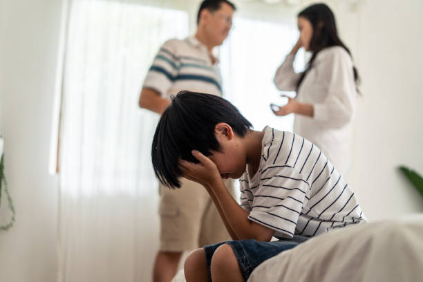 アジアの男の子の子供は、両親が自宅で戦ったり喧嘩したりしている間、ベッドに座って泣いています。顔や目を手で覆う子供は、暴力を見たくありません。家庭内の問題。 - 戦う ストックフォトと画像