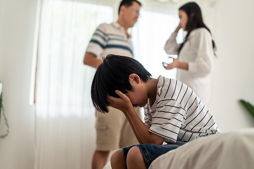 Asian boy kid sitting and crying on bed while parents having fighting or quarrel conflict at home. Child covering face and eyes with hands do not want to see the violence. Domestic problem in family.