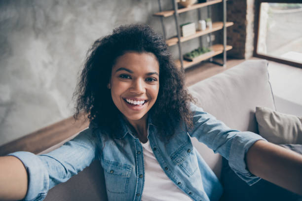 Closeup photo of pretty funny dark skin curly blogger lady making selfies excited toothy smiling social network blogger comfy couch casual denim outfit living room indoors Closeup photo of pretty funny dark skin curly blogger lady making, selfies excited toothy smiling social network blogger comfy couch casual denim outfit living room indoors self portrait stock pictures, royalty-free photos & images