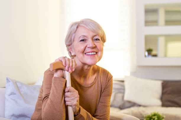 ソファに座っている笑顔の祖母。自宅の明るい背景に杖を歩いている美しい笑顔の先輩女性の肖像画。手を手で手をついて座っている老婦人 - human hand aging process senior adult cane ストックフォトと画像