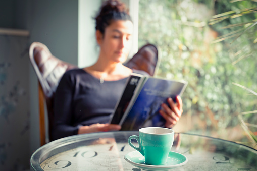 Hispanic woman at home sitting on modern chair in front of window relaxing in her living room reading a magazine and drinking tea, selective sharpness.