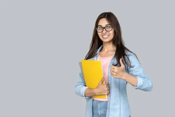 Cheerful and smiling young adult indian student teen girl holding copybooks in hand and showing thumb up. Happy woman standing isolated on grey background with copy space.