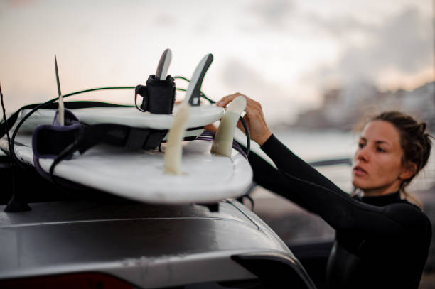 Surfer girl in black swimsuit taking surf board from silver car roof