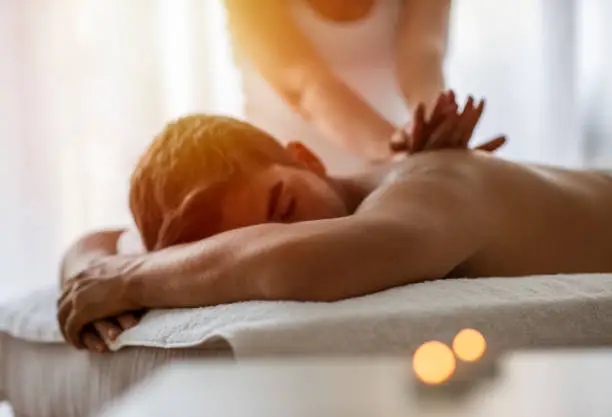 Masseur doing back massage on man body in the spa salon. Beauty treatment concept. Girl in a T-shirt doing a massage to a guy. Candles in the foreground. Man lying on the table on a white background.