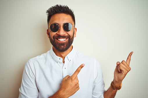Handsome indian buinessman wearing shirt and sunglasses over isolated white background smiling and looking at the camera pointing with two hands and fingers to the side.