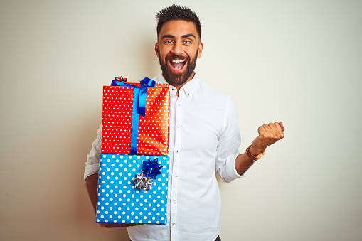 Young arab indian hispanic man holding birthday gifts standing over isolated white background screaming proud and celebrating victory and success very excited, cheering emotion