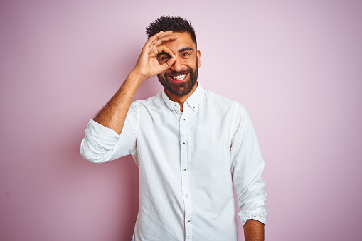 Young indian businessman wearing elegant shirt standing over isolated pink background doing ok gesture with hand smiling, eye looking through fingers with happy face.