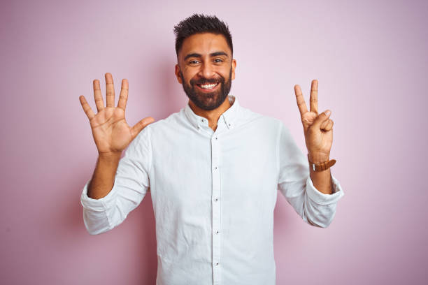 junge indische geschäftsmann trägt elegantes hemd stehen duponierend rosa hintergrund zeigt und zeigt mit den fingern nummer sieben, während lächelnd zuversichtlich und glücklich. - hispanic male stock-fotos und bilder