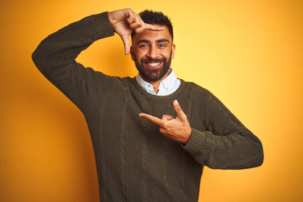 istock Young indian man wearing green sweater and shirt standing over isolated yellow background smiling making frame with hands and fingers with happy face. Creativity and photography concept. 1205720607