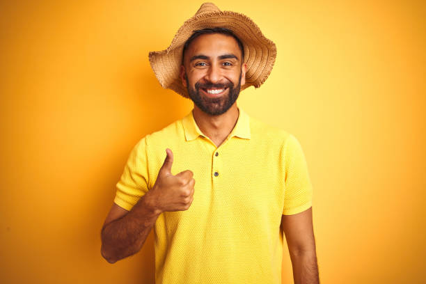 istock Young indian man on vacation wearing summer hat standing over isolated yellow background doing happy thumbs up gesture with hand. Approving expression looking at the camera with showing success. 1205720956