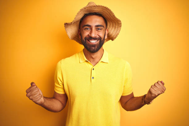 istock Young indian man on vacation wearing summer hat standing over isolated yellow background celebrating surprised and amazed for success with arms raised and open eyes. Winner concept. 1205720964