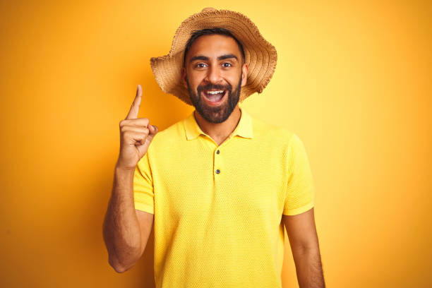 istock Young indian man on vacation wearing summer hat standing over isolated yellow background pointing finger up with successful idea. Exited and happy. Number one. 1205720993