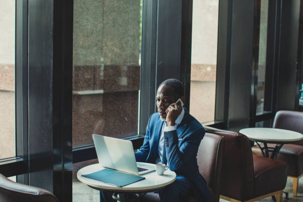 African Businessman Talking on his Cell Phone and looking at Laptop Elegant African businessman talking on his mobile phone while working on a laptop at modern cafe. businessman african descent on the phone business person stock pictures, royalty-free photos & images