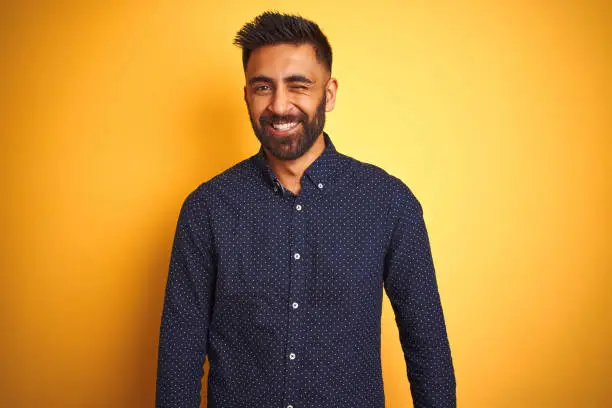 Young handsome indian businessman wearing shirt over isolated yellow background winking looking at the camera with sexy expression, cheerful and happy face.