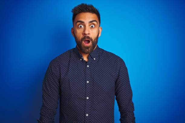 istock Young indian elegant man wearing shirt standing over isolated blue background afraid and shocked with surprise expression, fear and excited face. 1205722010