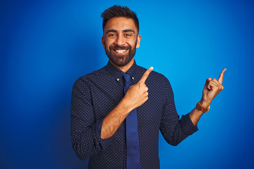 Young indian businessman wearing elegant shirt and tie standing over isolated blue background smiling and looking at the camera pointing with two hands and fingers to the side.