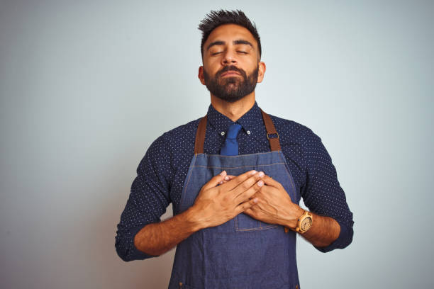 Young indian employee man wearing apron uniform standing over isolated white background smiling with hands on chest with closed eyes and grateful gesture on face. Health concept.