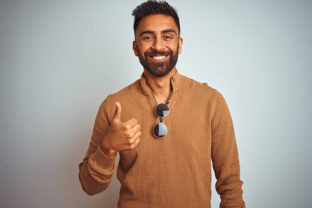 istock Indian man wearing elegant sweater and sunglasses standing over isolated white background doing happy thumbs up gesture with hand. Approving expression looking at the camera with showing success. 1205722865