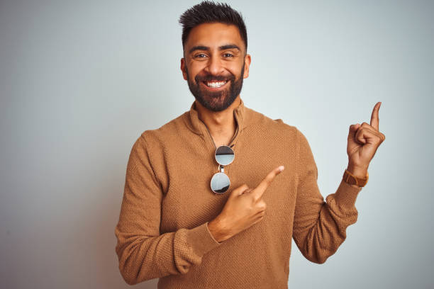 istock Indian man wearing elegant sweater and sunglasses standing over isolated white background smiling and looking at the camera pointing with two hands and fingers to the side. 1205722876