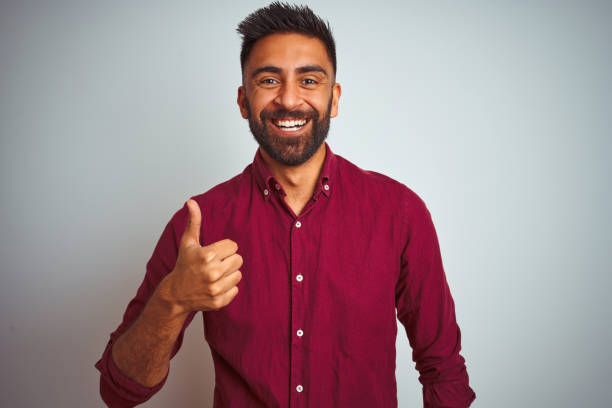 istock Young indian man wearing red elegant shirt standing over isolated grey background doing happy thumbs up gesture with hand. Approving expression looking at the camera showing success. 1205723229