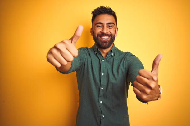 고립 된 흰색 배경 위에 서있는 우아한 셔츠를 입은 젊은 인도 사업가는 손으로 긍정적 인 제스처를 하고 엄지 손가락을 위로 하고 성공을 위해 행복합니다. 우승자 제스처. - human thumb cheerful human finger happiness 뉴스 사진 이미지