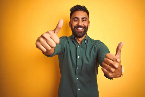 Young indian businessman wearing elegant shirt standing over isolated white background approving doing positive gesture with hand, thumbs up smiling and happy for success. Winner gesture.