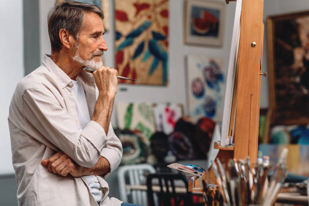 istock Side view of a thoughtful artist holding paintbrush while sitting in his studio. Male painter looking on canvas. 1205727016