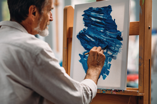 Close up of a pensive mature painter holds a paintbrush looking at a picture on the easel