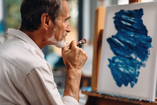 Close up of a pensive mature painter holds a paintbrush looking at a picture on the easel