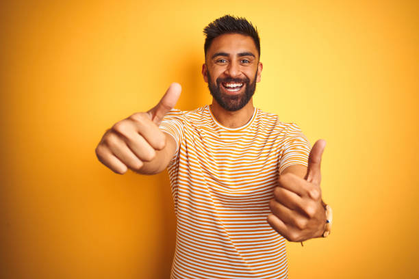 istock Young indian man wearing t-shirt standing over isolated yellow background approving doing positive gesture with hand, thumbs up smiling and happy for success. Winner gesture. 1205727406