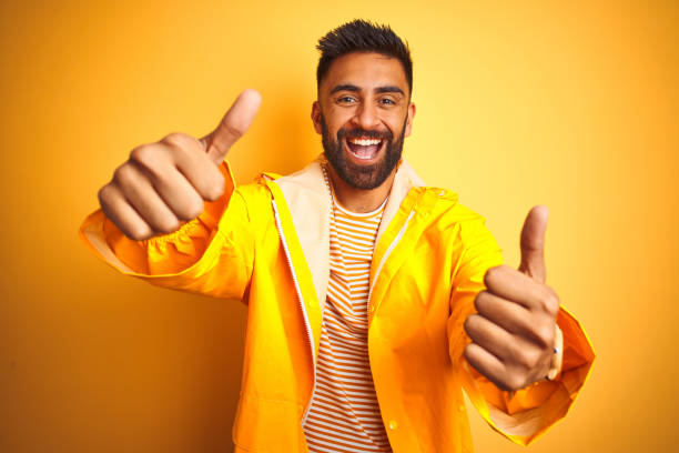 istock Young indian man wearing raincoat standing over isolated yellow background approving doing positive gesture with hand, thumbs up smiling and happy for success. Winner gesture. 1205730747