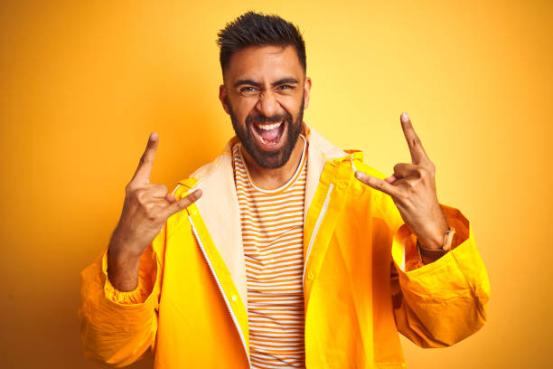istock Young indian man wearing raincoat standing over isolated yellow background shouting with crazy expression doing rock symbol with hands up. Music star. Heavy concept. 1205730782