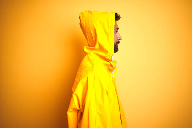 jovem indiano vestindo capa de chuva com capuz de pé sobre fundo amarelo isolado olhando para o lado, relaxa perfil pose com rosto natural com sorriso confiante. - hood men portrait profile - fotografias e filmes do acervo