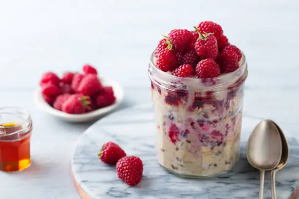 Overnight oats, bircher muesli with fresh raspberries in a glass jars. Marble cutting board. Close up.