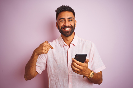 Joven indio usando teléfono inteligente de pie sobre fondo rosa aislado con la cara sorpresa apuntando con el dedo a sí mismo photo