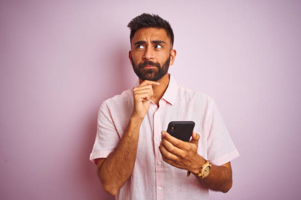 孤立したピンクの背景の上に立ってスマートフォンを使用して若いインドの男は、質問について考える深刻な顔、非常に混乱したアイデア - chin ストックフォトと画像