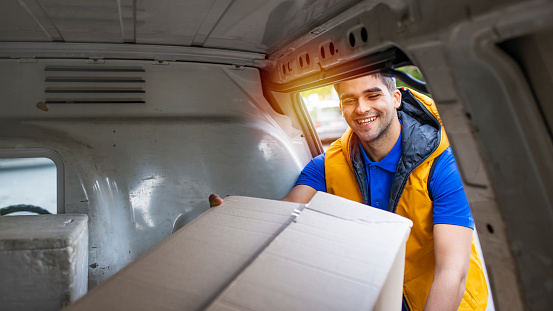 Proud delivery man in blue uniform holding parcel. Smiling courier standing in front of cargo van delivering package. Portrait of delivery man holding card box. View from the car trunk