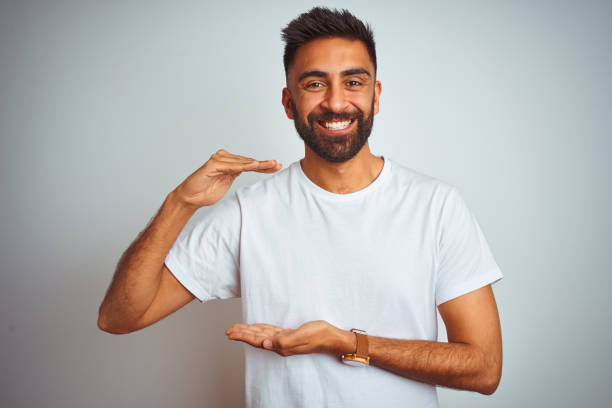 istock Young indian man wearing t-shirt standing over isolated white background gesturing with hands showing big and large size sign, measure symbol. Smiling looking at the camera. Measuring concept. 1205736456