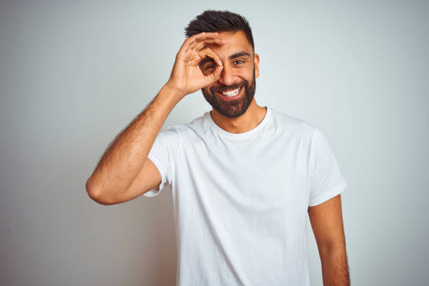 istock Young indian man wearing t-shirt standing over isolated white background doing ok gesture with hand smiling, eye looking through fingers with happy face. 1205736469