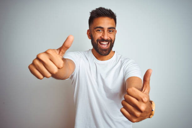 istock Young indian man wearing t-shirt standing over isolated white background approving doing positive gesture with hand, thumbs up smiling and happy for success. Winner gesture. 1205736485