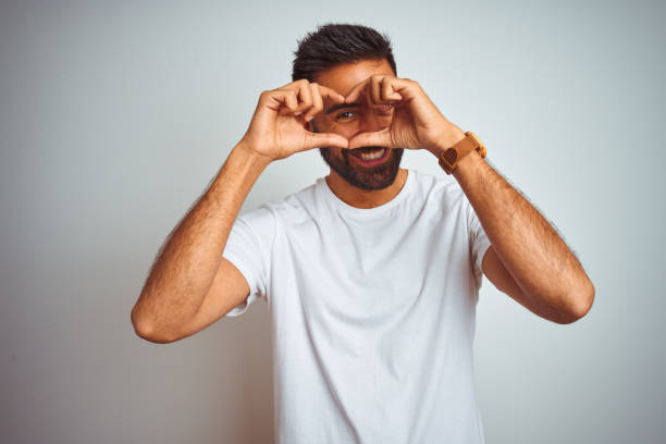 孤立した白い背景の上に立っているtシャツを着た若いインド人男性は、手と指で心臓の形をして看板を見て微笑む - isolated white men looking ストックフォトと画像