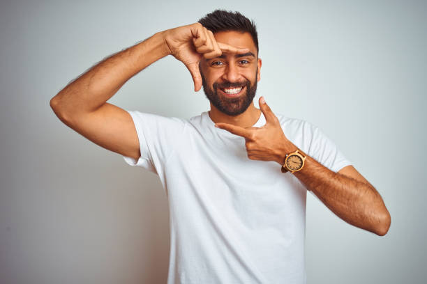 istock Young indian man wearing t-shirt standing over isolated white background smiling making frame with hands and fingers with happy face. Creativity and photography concept. 1205736491